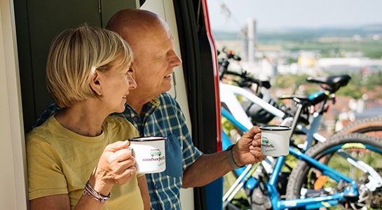 Elderly couple sitting in a campervan, drinking out of coffee mugs and overlooking the landscape.