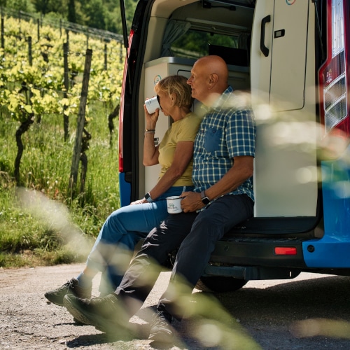 elderly couple in back of campervan next to a vineyard enjoying a cup of coffee