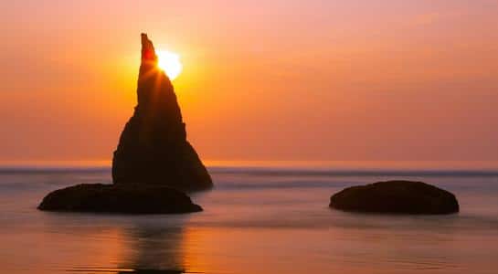 Sun setting behind Face Rock sea stack at Bandon Beach, Oregon, with waves washing ashore.