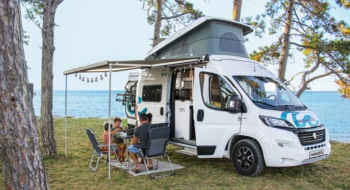 Family of four sitting around a camping table in front of their boxvan, parked at the seaside.