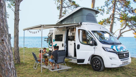 Family of four sitting around a camping table in front of their boxvan, parked at the seaside.