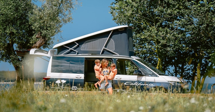 Mother with two children in her arms, smiling, standing in front of a roadsurfer Volkswagen California near a field of flowers