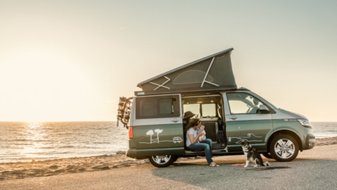 woman with a baby sitting inside the van, dog in front, sea and sunset in the background