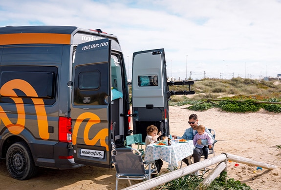 Father and his two children sitting at a camping table that is situated behind a parked boxvan on the beach.