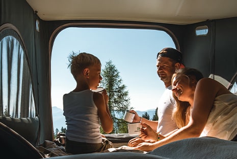 Family sitting in a roof top tent drinking out of mugs and looking at each other while tent is overlooking a blue sky and trees.