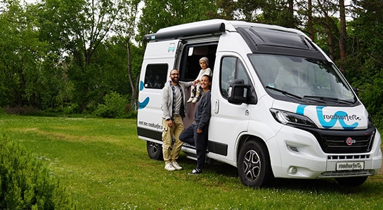 Family of three standing in between the sliding doors of a boxvan on a meadow