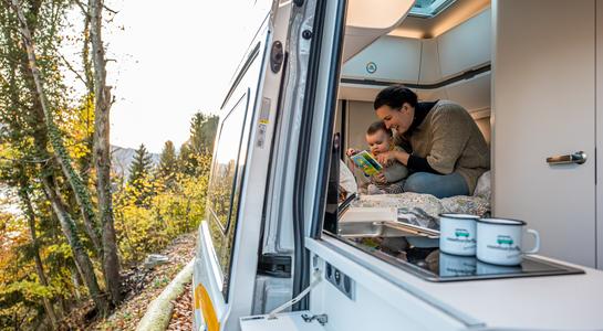 Mother and child reading a book together inside an RV parked by a forest
