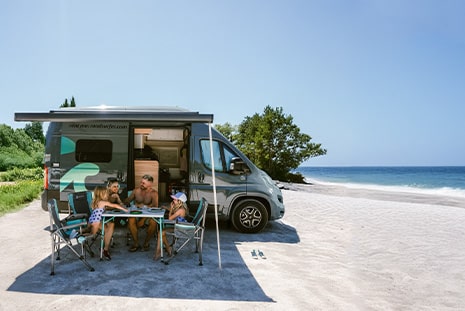 A family of four sitting at a table under the awning of a camper van, parked on a sunny beach with clear blue skies and ocean in the background.