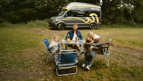 A family with children having breakfast at their campsite with their camper in a forest.