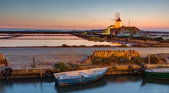 Rowing boats tied to the dock of a farmland during the evening. You can see an illuminated windmill in the back of the picture.