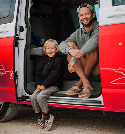 Father and son smiling and sitting in the doorway of a red campervan