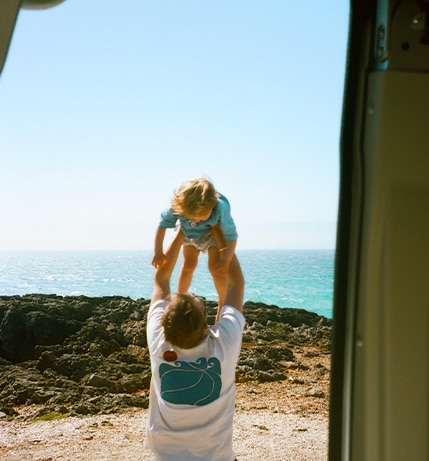 joyful moment of a man lifting a young child into the air near a rocky coastline, with the ocean and clear blue sky in the background