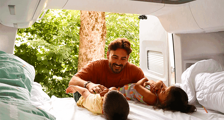 A father playing with his two young children on a bed inside a camper van, smiling and laughing together. Sunlight streams in through the open rear doors, revealing lush green trees outside and creating a warm, family-friendly travel atmosphere.