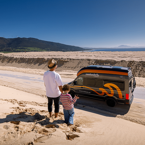 A father holding his son's hand as they descend a sand dune in southern Spain toward their campervan.