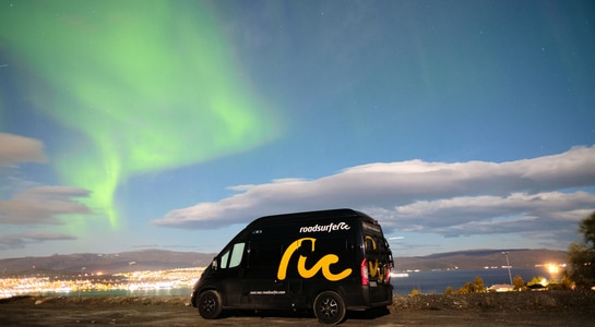 Black and yellow Fiar Ducato camper standing with views to a port city and northern lights in the sky