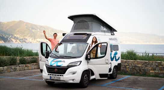 Couple standing on the doorstep of a Fiat Ducato camper right by the sea in Italy