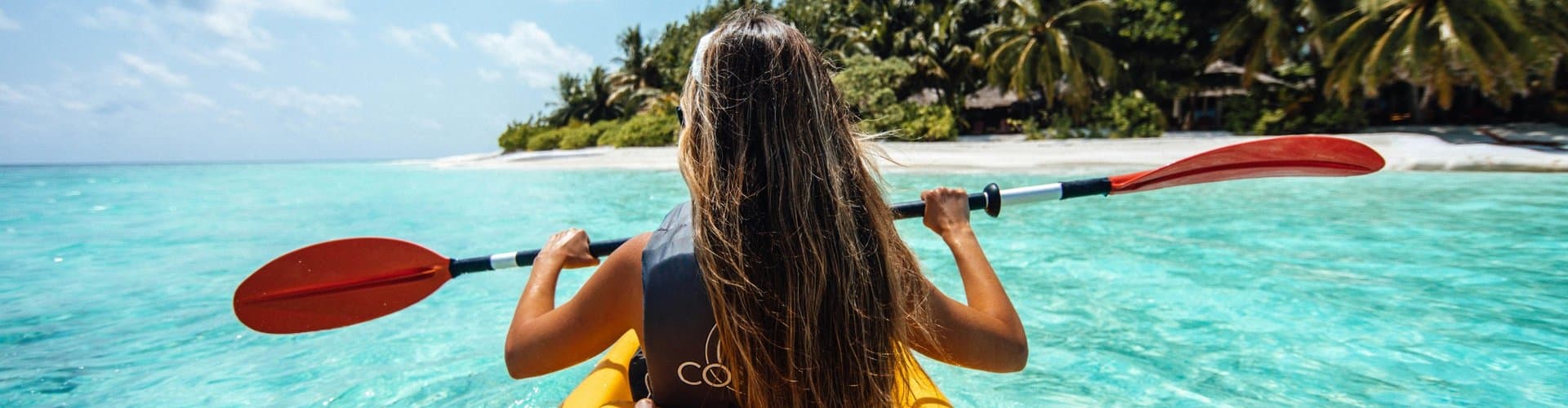 A person kayaking toward a palm-lined beach in clear turquoise water under a bright blue sky