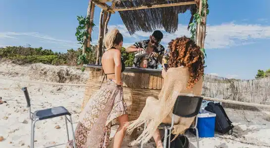 Bartender mixing tropical drinks for two women at a rustic tiki bar on a sandy beach under a clear blue sky in the Florida Keys.