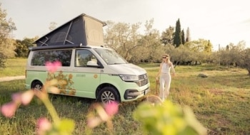 Roadsurfer VW camper with flower design stands with open pop-up roof on a field while a young woman walks her dog.