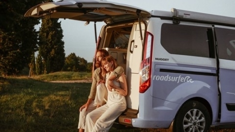 Couple sitting in the trunk of a blue Ford Nugget campervan from roadsurfer and looking towards the sunshine