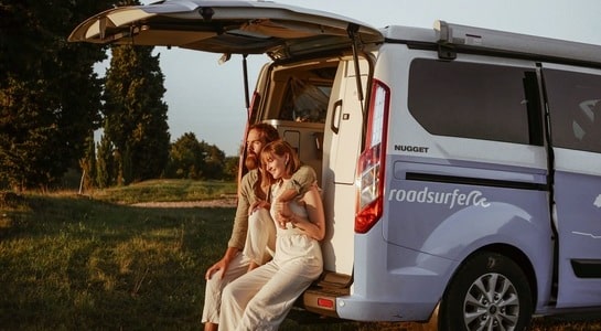Couple sitting in the trunk of a blue Ford Nugget campervan from roadsurfer and looking towards the sunshine