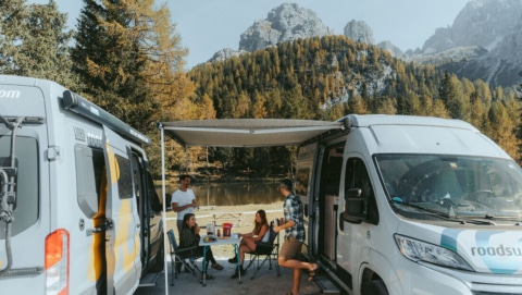 Two parked campers with a mountain backdrop and a group of friends sitting under the awnings of the campers and having fun