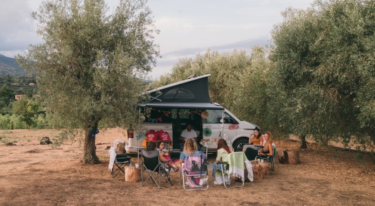 Group of friends sitting on camping chairs in a circle in front of a campervan parked in an olive grove
