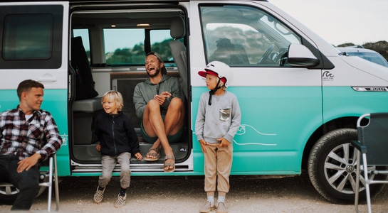 Two family friends with their two children sit in a turquoise roadsurfer campervan and laugh.
