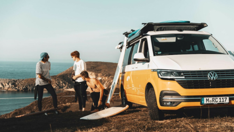 three friends with surfboards and a yellow campervan at the ocean