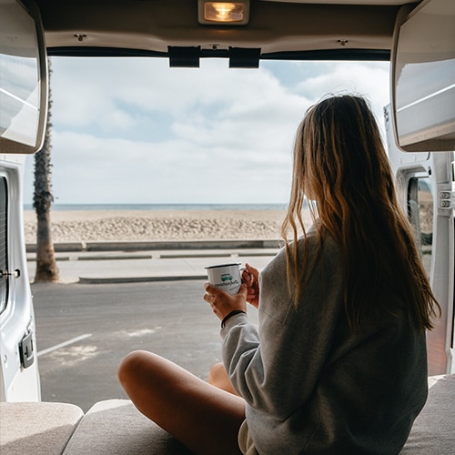 Girl sitting in the back of a roadsurfer camper van drinking a coffee, looking at the beach