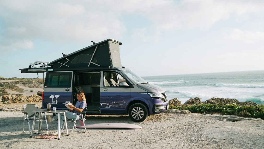 Girld reading a book in front of a campervan at the coast