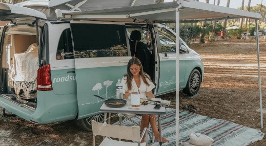 Girl in white dress sitting next to a picnic table in front of a roadsurfer camper in summer