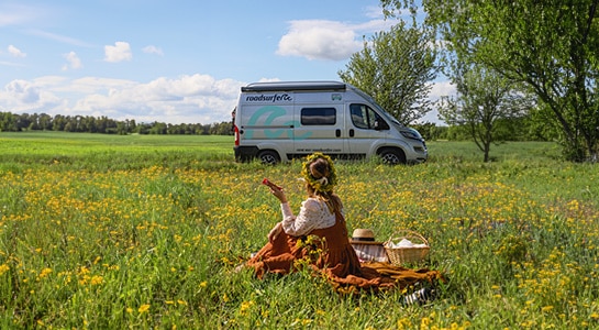 A girl with a flower crown sits in a field with a roadsurfer camper in the background