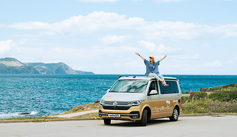 Joyous girl with arms stretched sitting on the roof of a VW campervan from roadsurfer