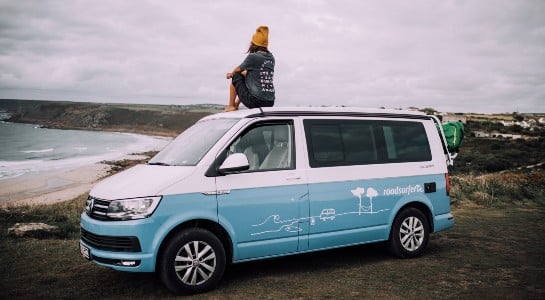 girl with a cap sitting on top of a campervan looking at the sea