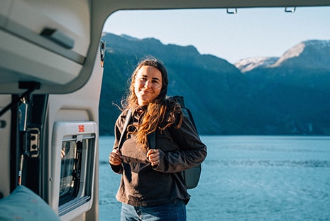 Girl with a backpack standing behind a camper in front of a lake in Sweden