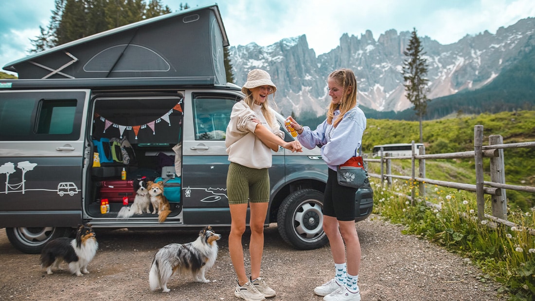 Two girls with their dogs and a campervan in front of mountains using Autan mosquito spray