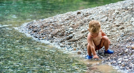 Toddler squatting by the clear American River on a pebbly shore—cheap things to do with kids in California.