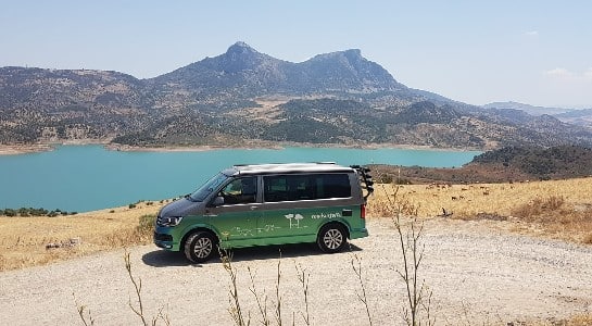Green and silver VW California standing in front of a lake with mountains in the background
