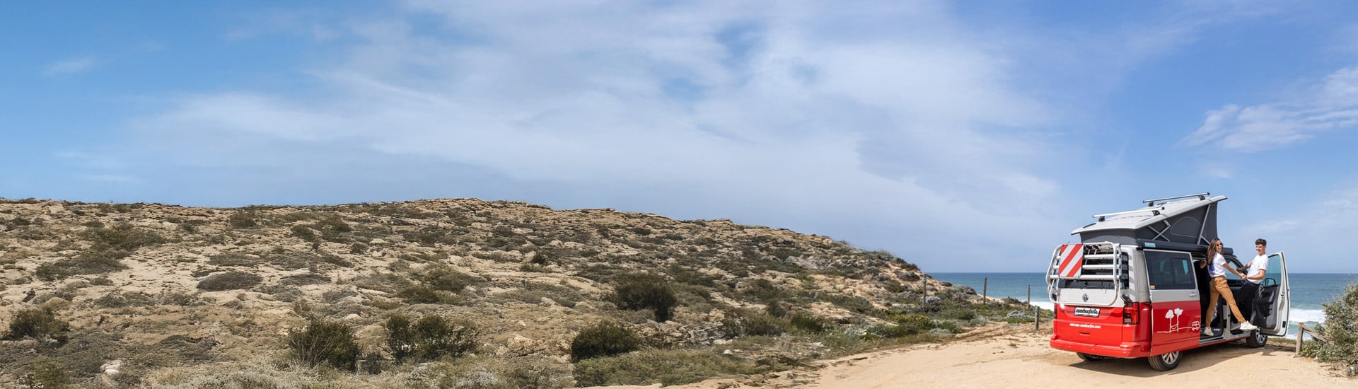 A couple enjoys a scenic beachside moment beside their red and white camper van with a pop-up roof, parked on a sandy cliff overlooking the ocean.