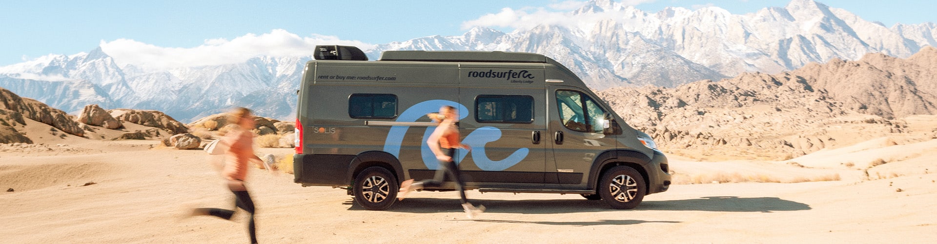 Two women running past a roadsurfer Liberty Lodge RV in the desert with snow-capped mountains in the background
