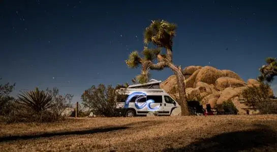 RV parked beside Joshua trees and boulders under a starry desert sky