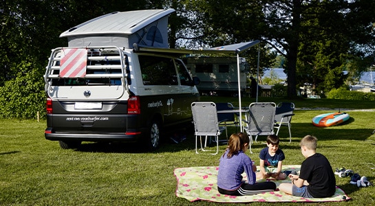 Children sitting on a picnic blanket in front of a dark grey campervan parked on a campsite close to the lake.