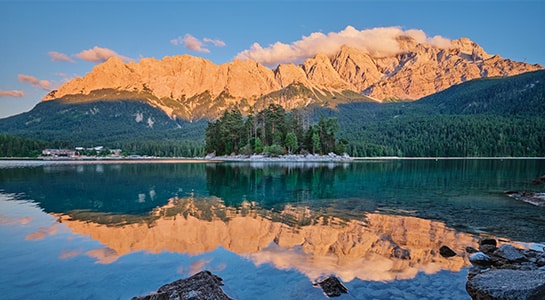 Maximiliansinsel island surrounded by the clear waters of Lake Eibsee, with the Zugspitze mountains bathed in warm evening light in the Bavarian Alps.