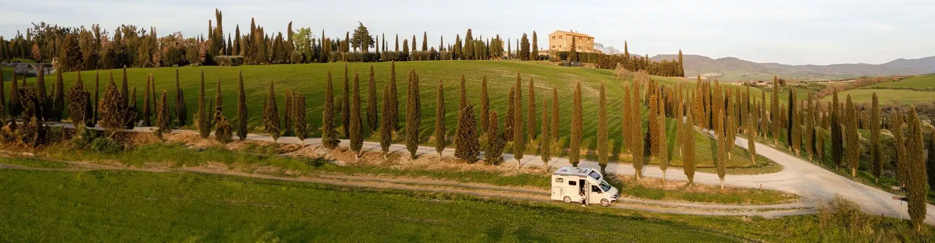 Landscape scenery over Tuscany. Green fields and tall green trees. White motorhome parked on a pathway in between the fields. Hills in the background.