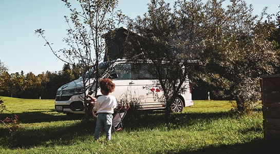 Little kid standing in a field in front of a campervan parked behind some smaller trees.