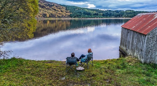 Travelers relaxing by a peaceful lake with autumn trees during a Scotland road trip near Loch Lomond