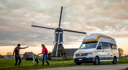 man and woman playing with dog in front of a windmill and a campervan