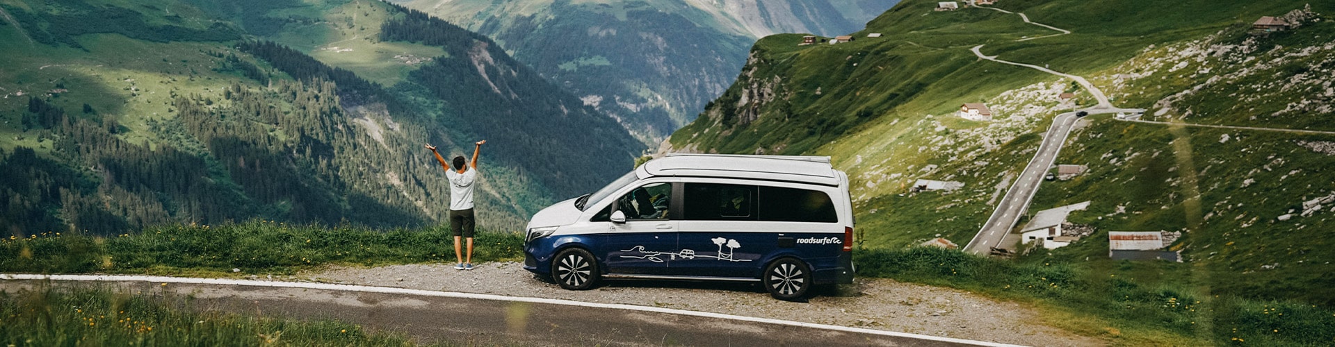 Man standing next to blue and white campervan, putting his arms into the sky and overlooking a valley in the mountains.