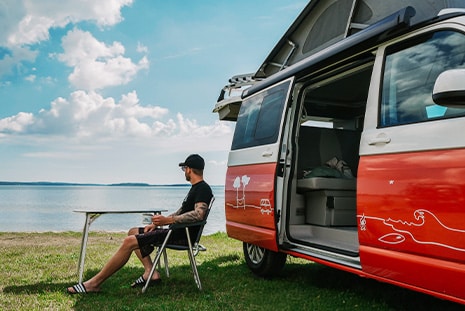 Man relaxing with a cup of coffee in a camping chair in front of a campervan enjoying the lake view